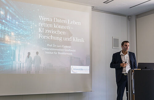 Prof. Dr. Lars Kaderali (Universitätsmedizin Greifswald) spricht zum Einsatz von KI in der Gesundheitsforschung Ein Mann hält einen Vortrag vor einem großen Bildschirm mit Text zu KI und Forschung