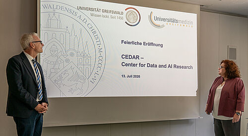 Prof. Dr. Karlhans Endlich und Prof. Dr. Katharina Riedel eröffnen das CEDAR Zwei Personen, ein mann und eine Frau, stehen vor einer Präsenation und halten eine Ansprache.