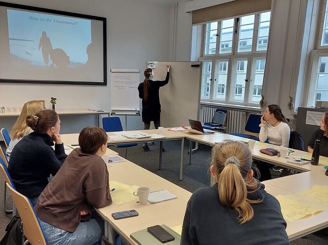 Lernende sitzen an einem o-förmig aufgebauten Konferenztisch, der Dozent schreibt etwas auf ein Whiteboard.
