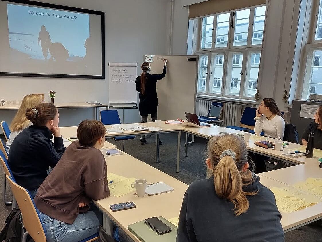 Frauen sitzen an o-förmigen Tisch bei einem Workshop während die Dozentin auf einem Whiteboard schreibt.