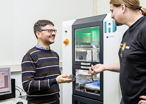 Professor Hermann Seitz (l.) und Dr. Vincent Morrison überprüfen ein 3D-gedrucktes Teil. Im Hintergrund eine Serienmaschine der Firma AIM3D 