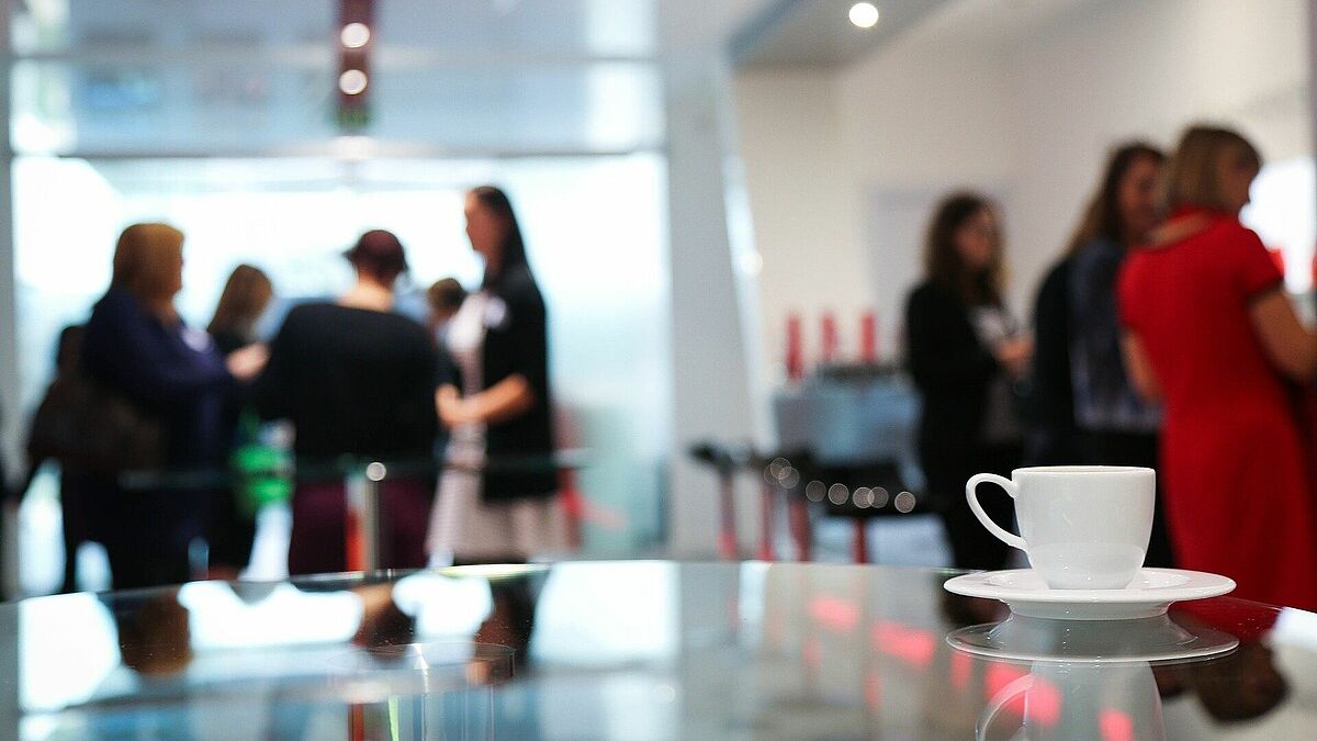 Eine weiße Kafeetasse steht auf einem Glastische. Im Hintergrund sind unschaft Frauen zu erkennen, die sich austauschen.