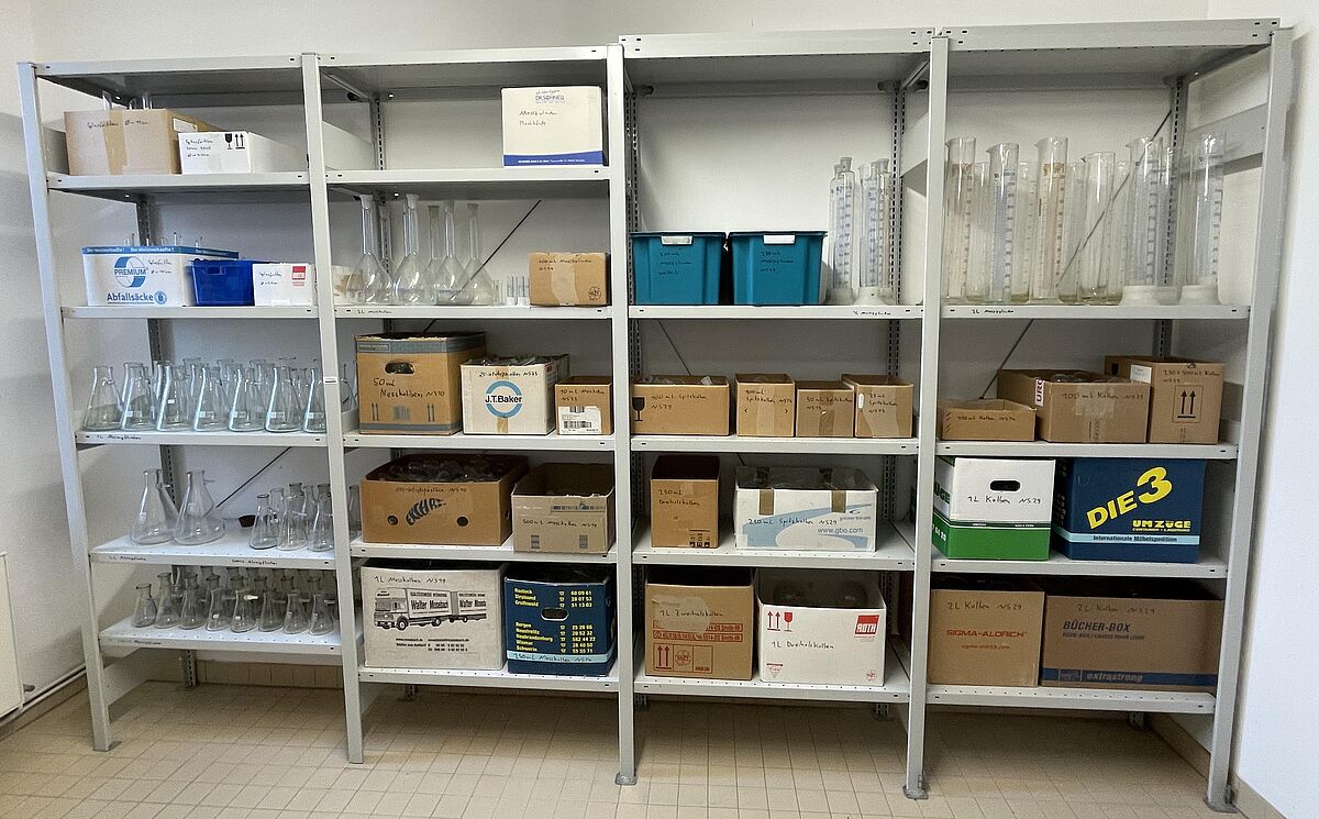 Ein Foto aus dem Glaslager im Entsorgungshof der Universität Rostock. Das Foto zeigt ein sehr breites und hohes Regal, welches mit zahlreichen Kisten und freistehenden Glaswaren gefüllt ist. Man sieht unter anderem Messzylinder, Kolben und Absaugflaschen.