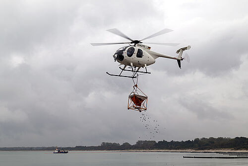 Die luftgestützte Binderausbringung durch einen Hubschrauber vom Typ Hughes 500 am Strand von Rostock Hohe Düne. Im Hintergrund zu sehen ist das Schiff „Flunder“, das von der Rostocker Firma BALTIC TAUCHER zur Ölabwehr eingesetzt wird. 